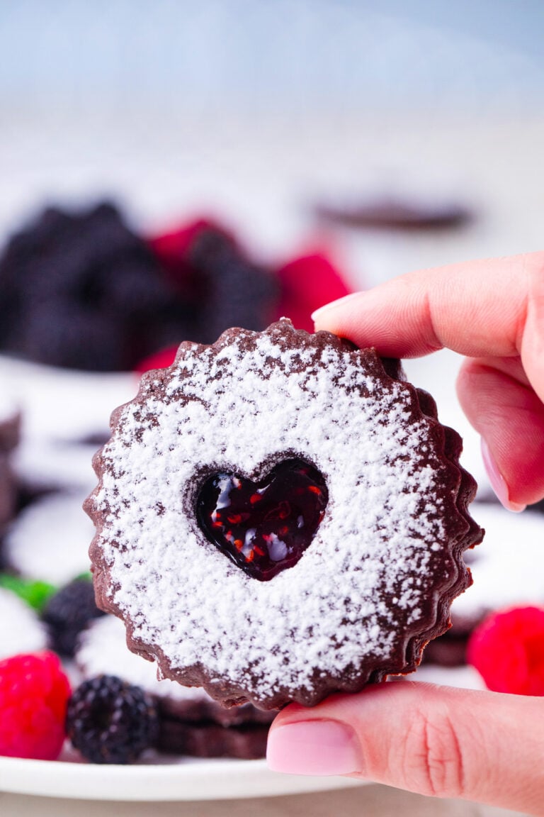 A hand holding a chocolate Linzer cookie filled with raspberry jam.