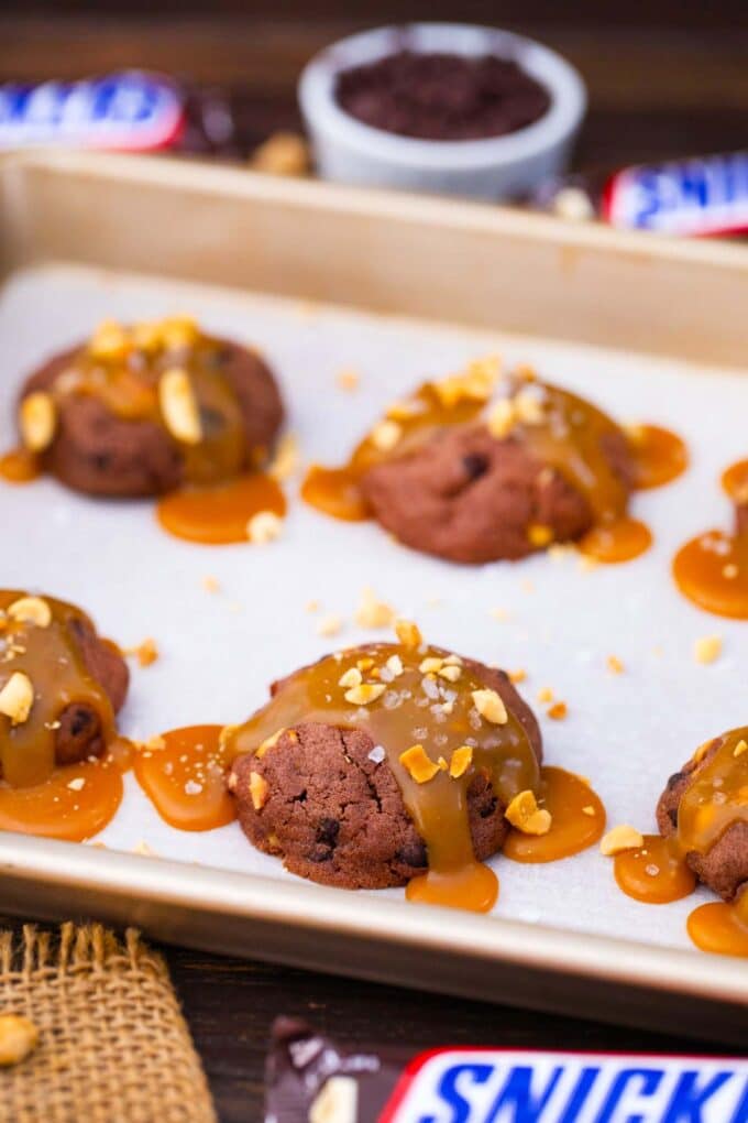 snickers cookies topped with caramel sauce and peanuts on a baking sheet