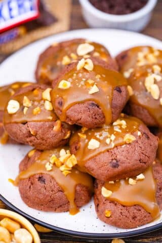 close shot of snickers cookies on a plate, topped with caramel sauce and peanuts