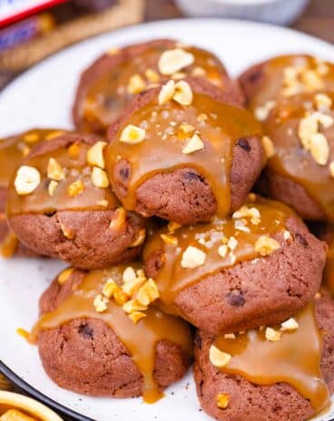 close shot of snickers cookies on a plate, topped with caramel sauce and peanuts