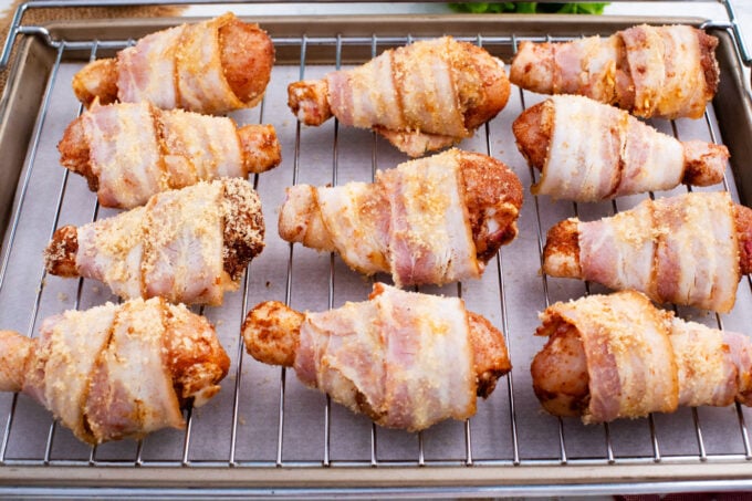 Brown sugar coated bacon wrapped chicken drumsticks on a wire rack over a baking sheet.