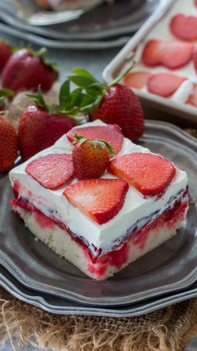 Sliced strawberry jello poke cake topped with whipped cream and sliced strawberries on a silver plate