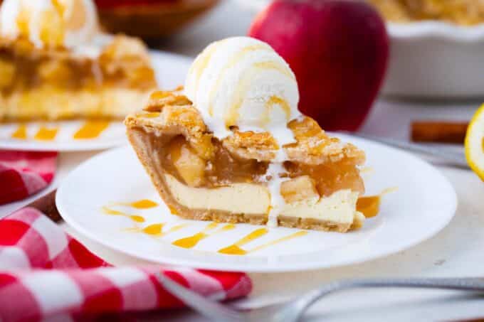 a plate with a slice of apple pie with cream cheese layer topped with ice cream, a red apple is in the background