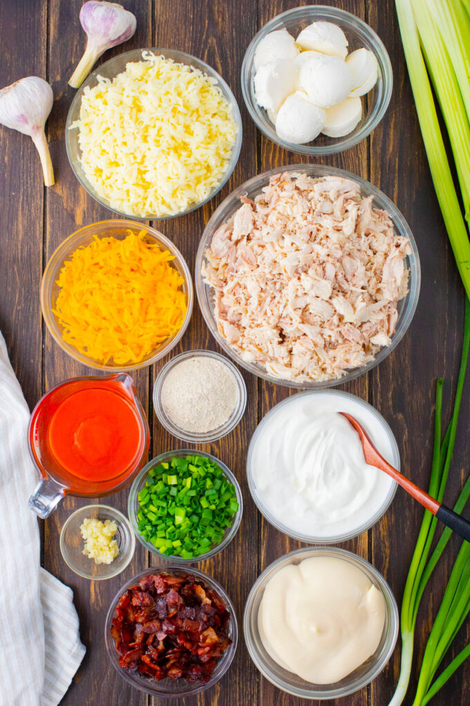 Overhead shot of Buffalo crack chicken dip ingredients in bowls on a wooden table.