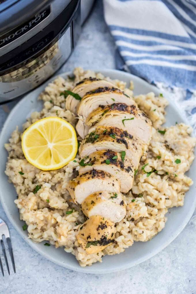 Sliced chicken breast with lemon over creamy brown rice on a plate with slow cooker in the background