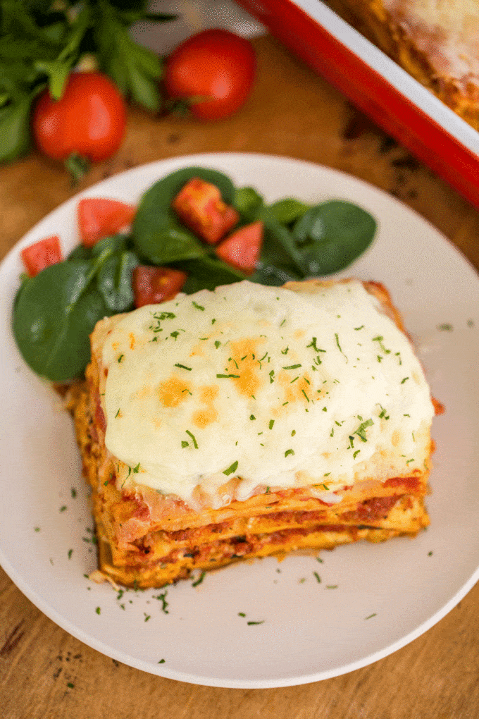 A slice of lasagna on a white plate next chopped tomatoes and fresh spinach