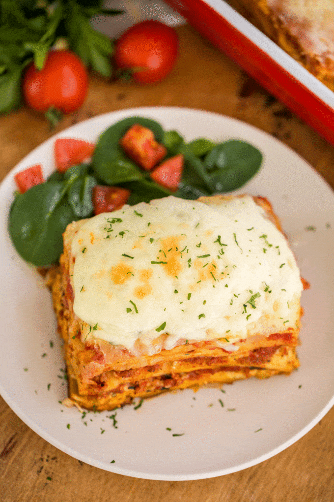 A slice of lasagna on a white plate next chopped tomatoes and fresh spinach 