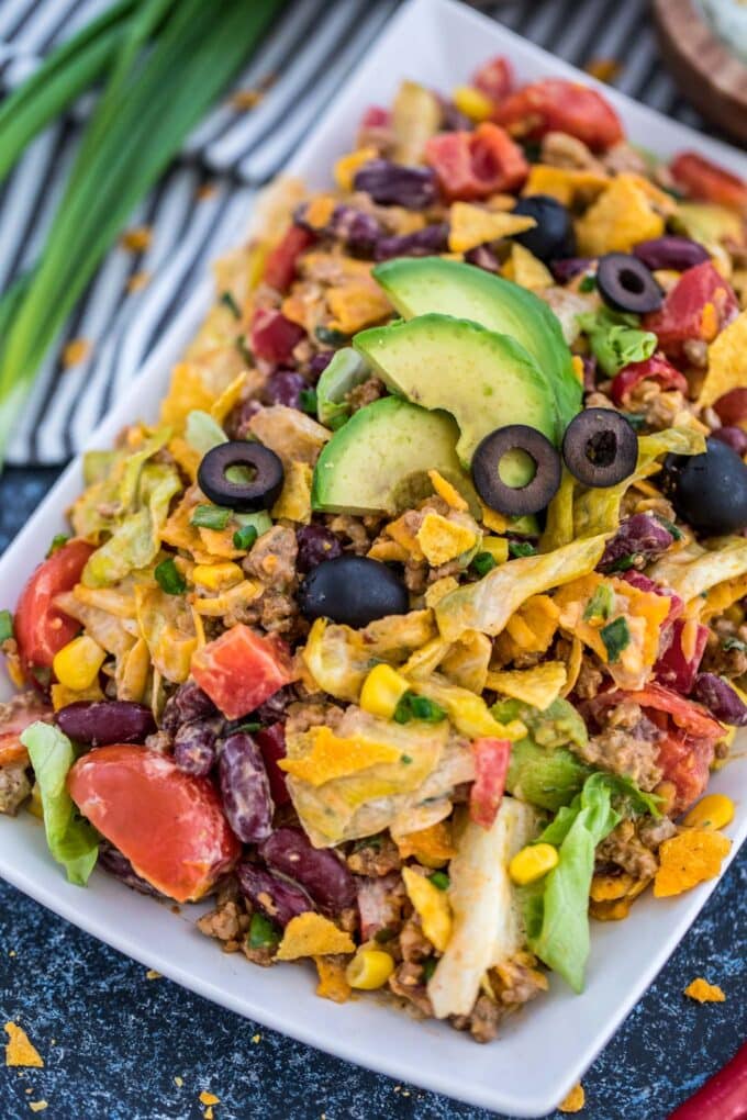 Taco salad with ground beef, avocado, tortilla chips, lettuce, beans, olives on a white plate.