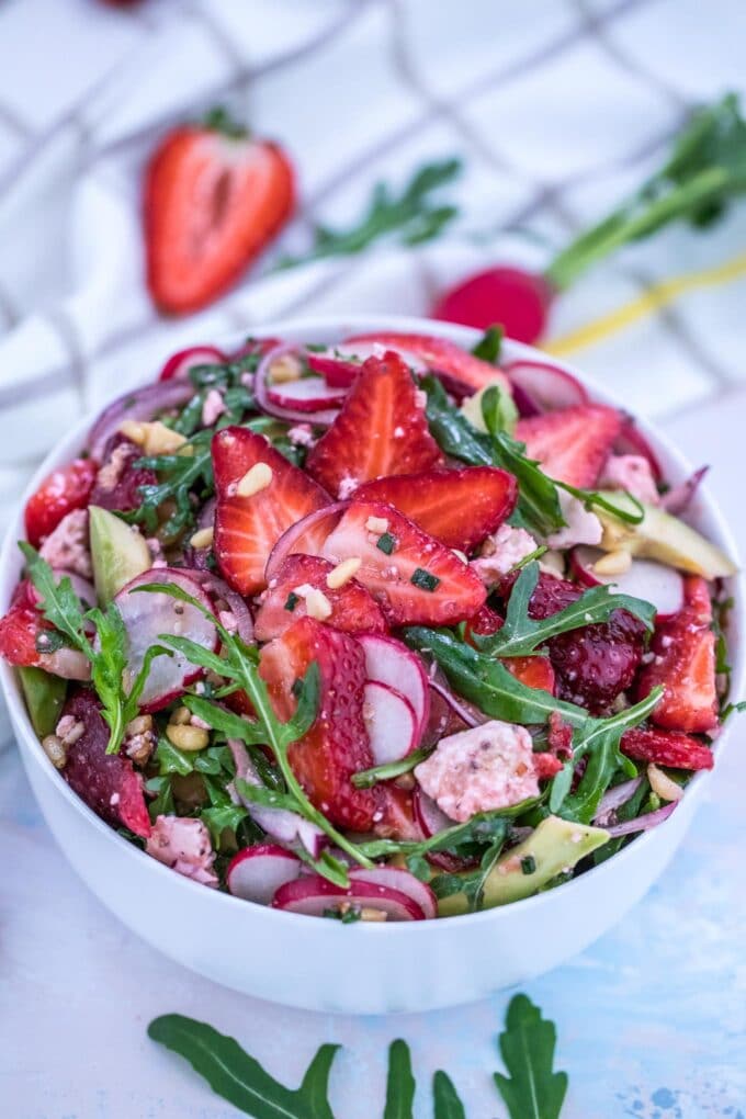 Strawberry Arugula salad with sliced strawberries, onion, avocado, radish, and feta cheese in a white bowl. 