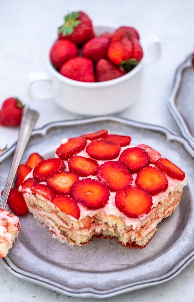 a slice of creamy strawberry tiramisu on a serving plate, with a bowl of fresh strawberries next to it