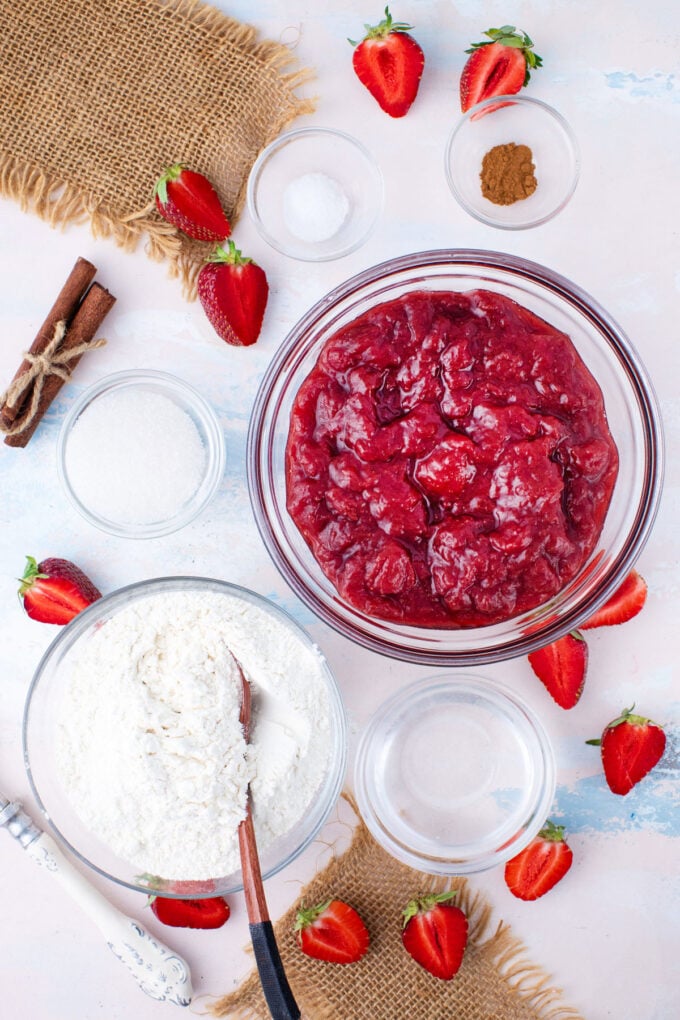 Overhead shot of strawberry pie ingredients in bowls on a light pink surface.
