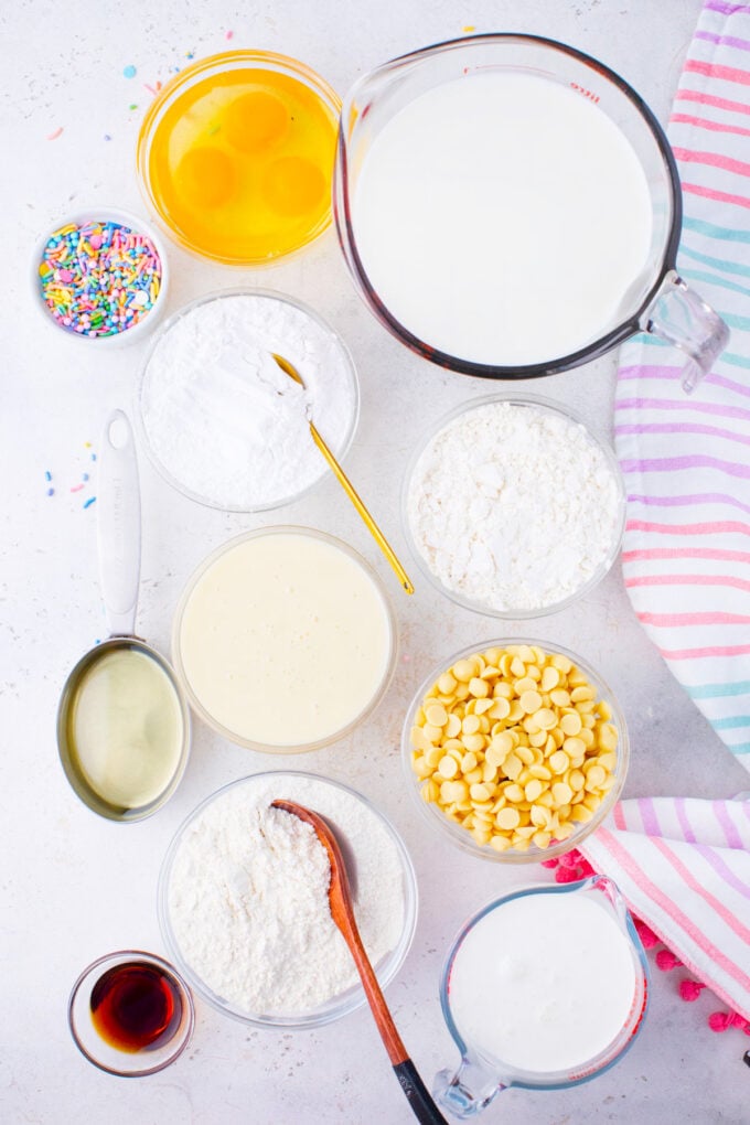 Overhead shot of unicorn poke cake ingredients in bowls on a white surface.