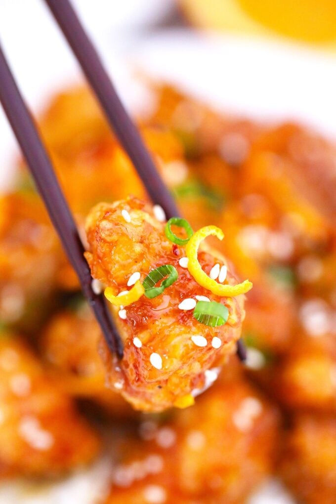 A slice of chicken marinated in orange sauce and garnished with sesame seeds and green onions on chop sticks. 