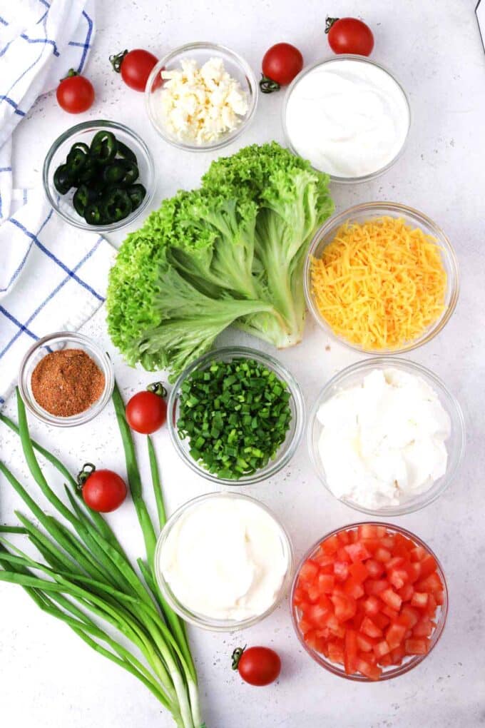 Overhead shot of taco dip ingredients in bowls on a white surface.