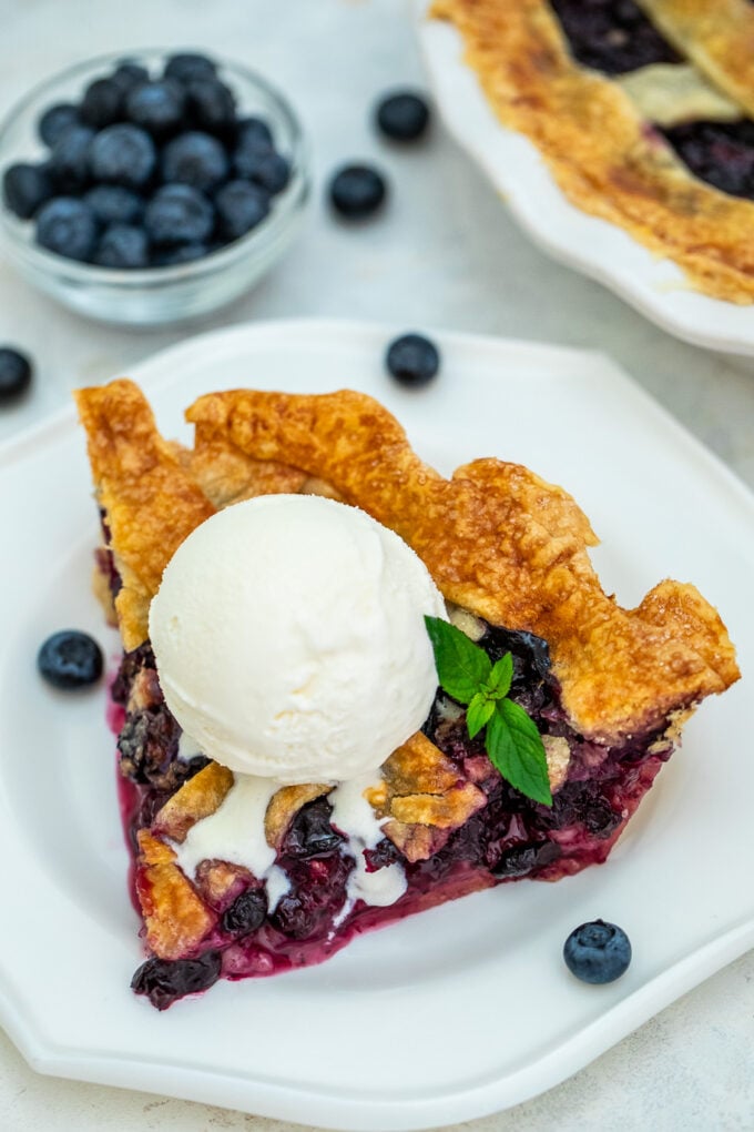 Golden blueberry pie slice topped with a dollop of ice cream and fresh blueberries in a bowl next to it