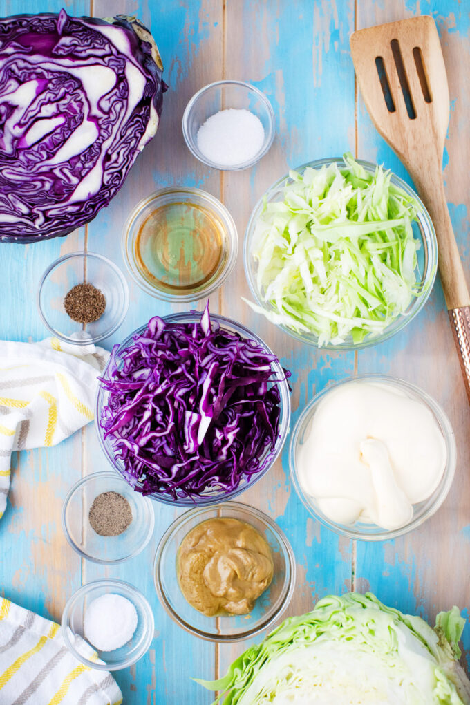 Overhead shot of coleslaw ingredients in bowls on a blue table.