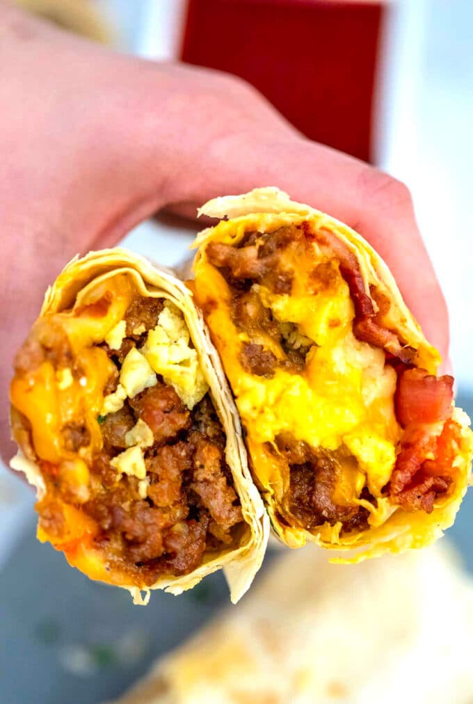 A hand holding two halves of sausage egg and cheese breakfast burrito oozing with cheese.