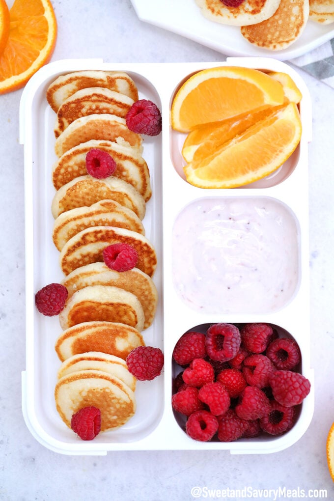 Photo of a lunch box with mini pancakes, yogurt, raspberries and oranges.