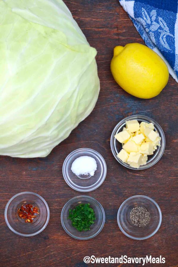 boiled cabbage ingredients: cabbage head, butter, salt, red pepper flakes, parsley, black pepper, and lemon on a wooden table