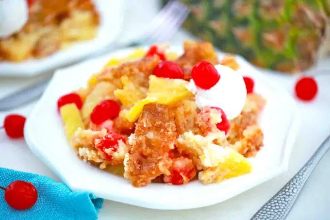 a plate of pineapple dump cake with maraschino cherries and whipped cream and a fresh pineapple in the background