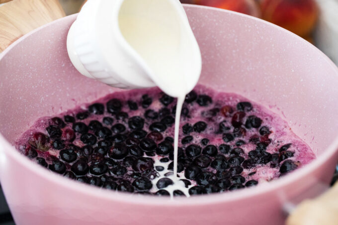 Pouring cornstarch slurry into blueberry pie filling in a saucepan.
