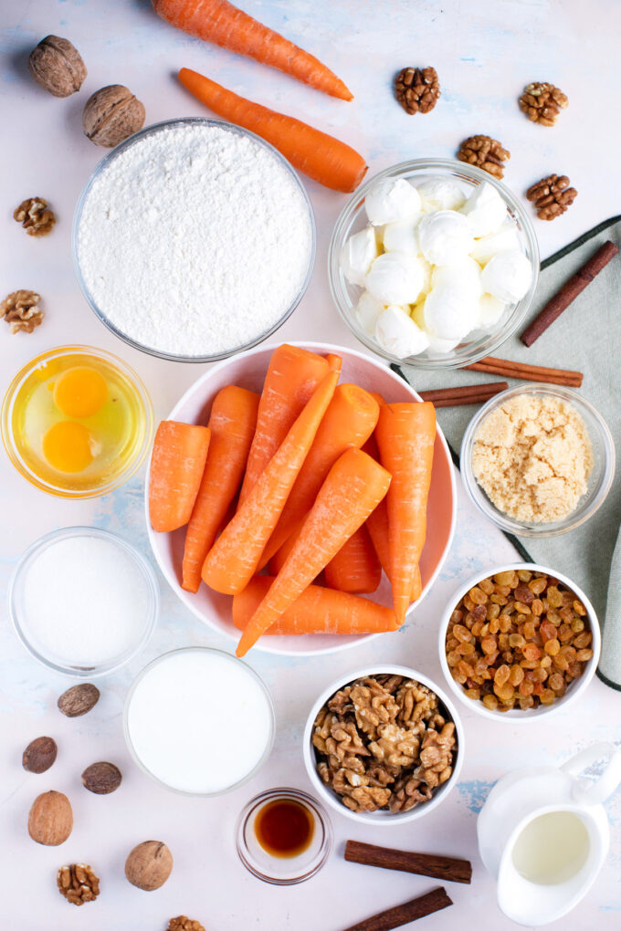 Overhead shot of carrot cake cheesecake ingredients in bowls on a white surface.