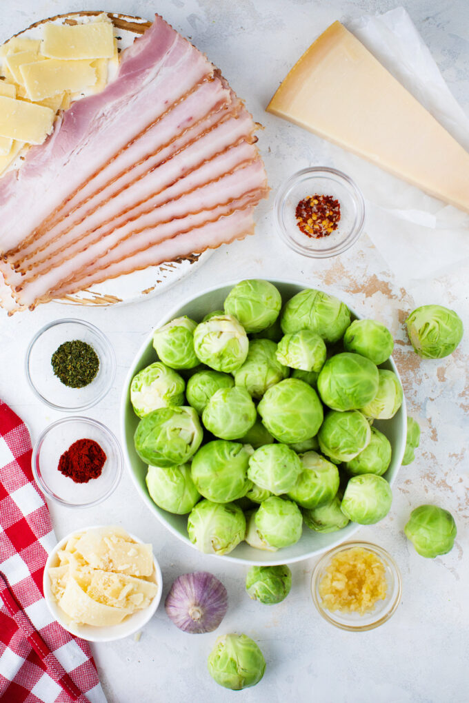Overhead shot of brussels sprouts with bacon ingredients in bowls on a white surface.