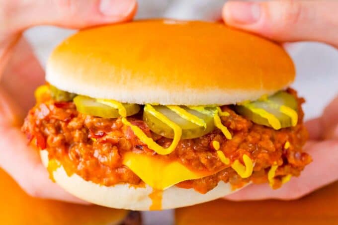 Hands holding bacon cheeseburger sloppy joes, drizzled with mustard and topped with pickles.