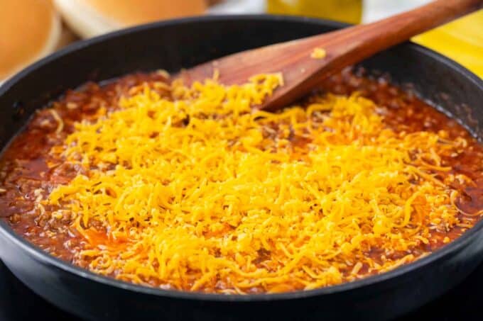Stirring shredded cheddar cheese into bacon cheeseburger sloppy joes meat mixture in a skillet.
