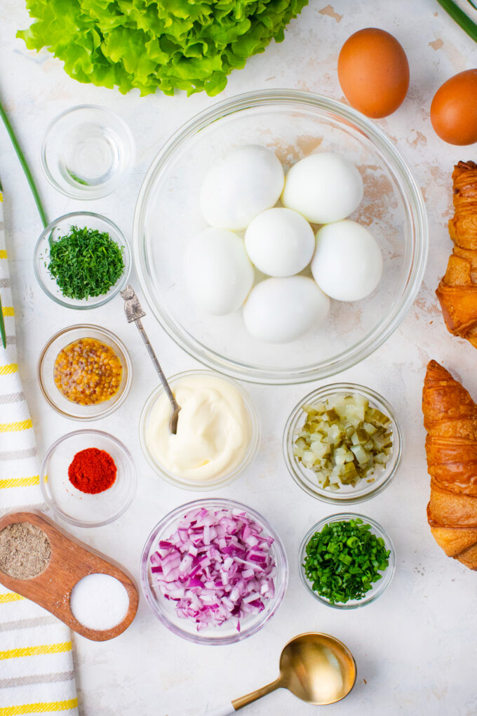 Overhead shot of classic egg salad ingredients in bowls on a white surface.