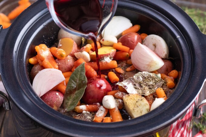 Adding red wine to a slow cooker with root vegetables and chuck roast.