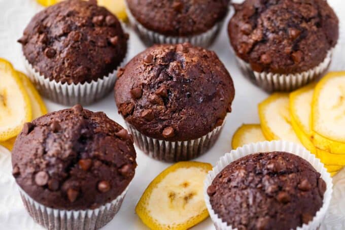 Chocolate banana muffins next to banana slices on a white surface.