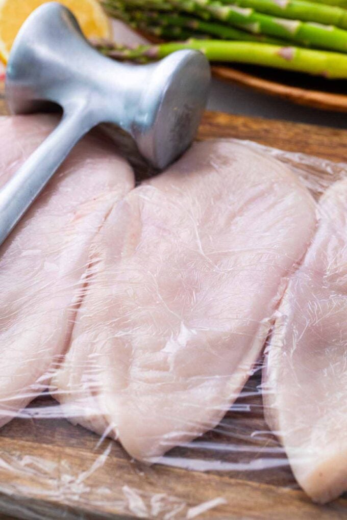 Chicken breasts covered with plastic wrap on a wooden board with a mallet next to it.