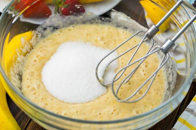 Adding sugar to a bowl with banana and egg mixture.
