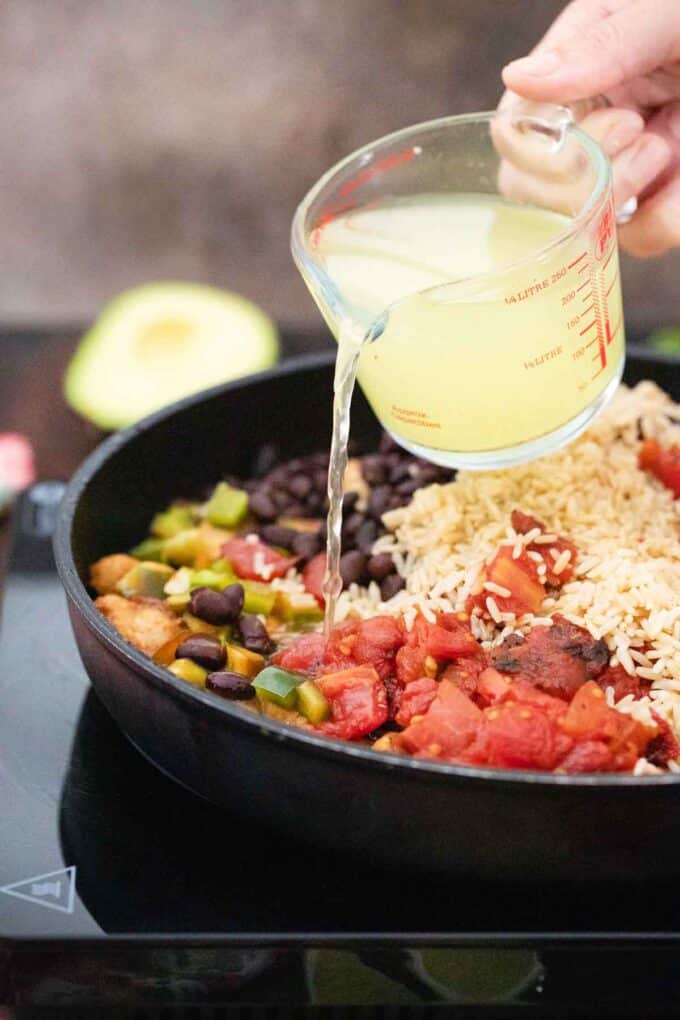 pouring broth into a pan with rice beans tomatoes and chicken
