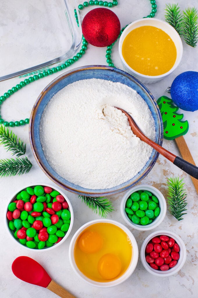 Overhead shot of M&M's cookie bars ingredients in bowls on a white surface.