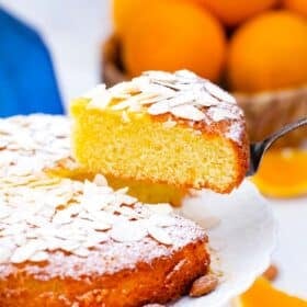 lifting a slice of almond orange cake from a cake stand and fresh oranges in the background