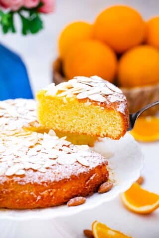 lifting a slice of almond orange cake from a cake stand and fresh oranges in the background