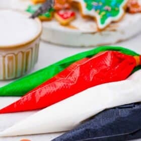 four piping bags filled with colorful royal icing: greed red white black on a table with cookies in the background