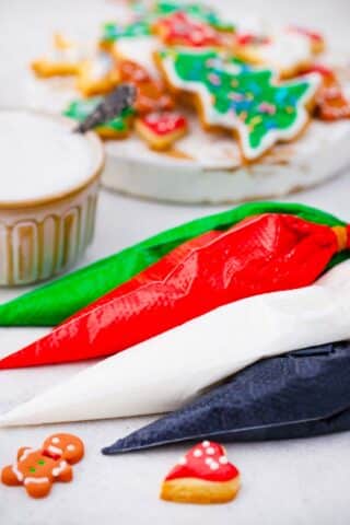 four piping bags filled with colorful royal icing: greed red white black on a table with cookies in the background
