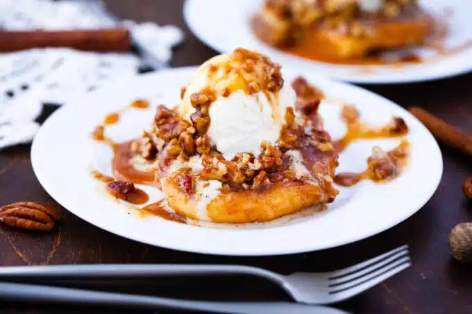 A dessert plate featuring a scoop of vanilla ice cream on top of a slice of baked apple, drizzled with caramel sauce and sprinkled with walnuts. A silver fork and knife rest beside the plate on a dark wooden table.