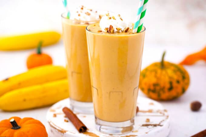 two tall glasses with creamy pumpkin pie smoothie topped with whipped cream and chopped pecans with banana and mini pumpkins in the background