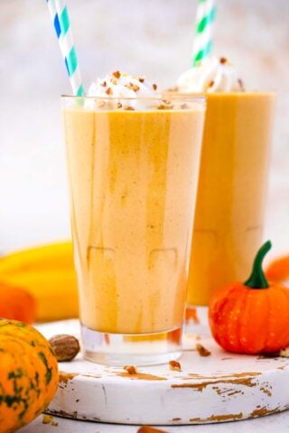 two glasses of pumpkin pie smoothie on a white cutting board with mini pumpkin next to it