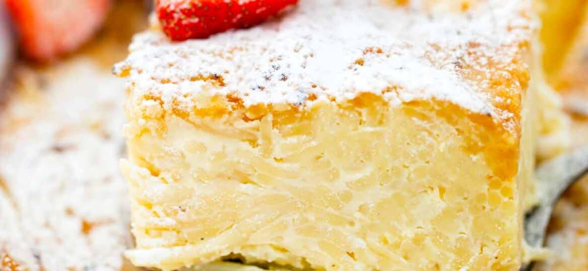a pie cutter with a slice of macaroni pudding on top of macaroni pudding casserole with powdered sugar and topped with sliced strawberries