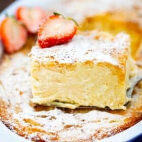 a pie cutter with a slice of macaroni pudding on top of macaroni pudding casserole with powdered sugar and topped with sliced strawberries