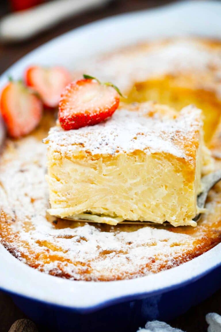 a pie cutter with a slice of macaroni pudding on top of macaroni pudding casserole with powdered sugar and topped with sliced strawberries