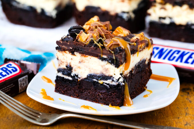 Slutty cheesecake brownie bar with brownie layer, cheesecake layer, Oreo layer, snickers, and chocolate ganache.