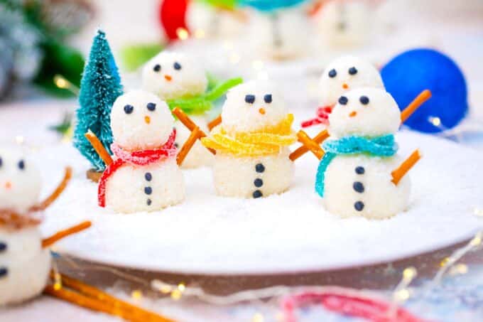 snowman truffles with different colors of candy saris and pretzel hands on a plate with shredded coconut
