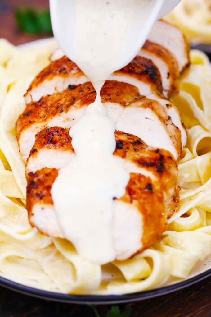 Pouring Alfredo sauce over sliced chicken breast on top of pasta.