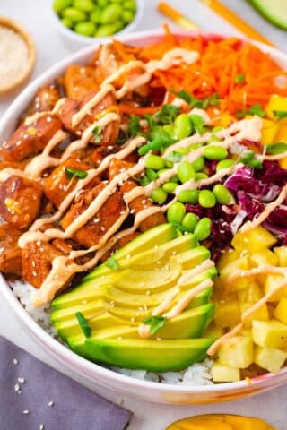 cooked salmon poke bowl with shredded red cabbage shredded carrot edamame pineapple mango and avocado with a spicy mayo drizzle and sesame seeds